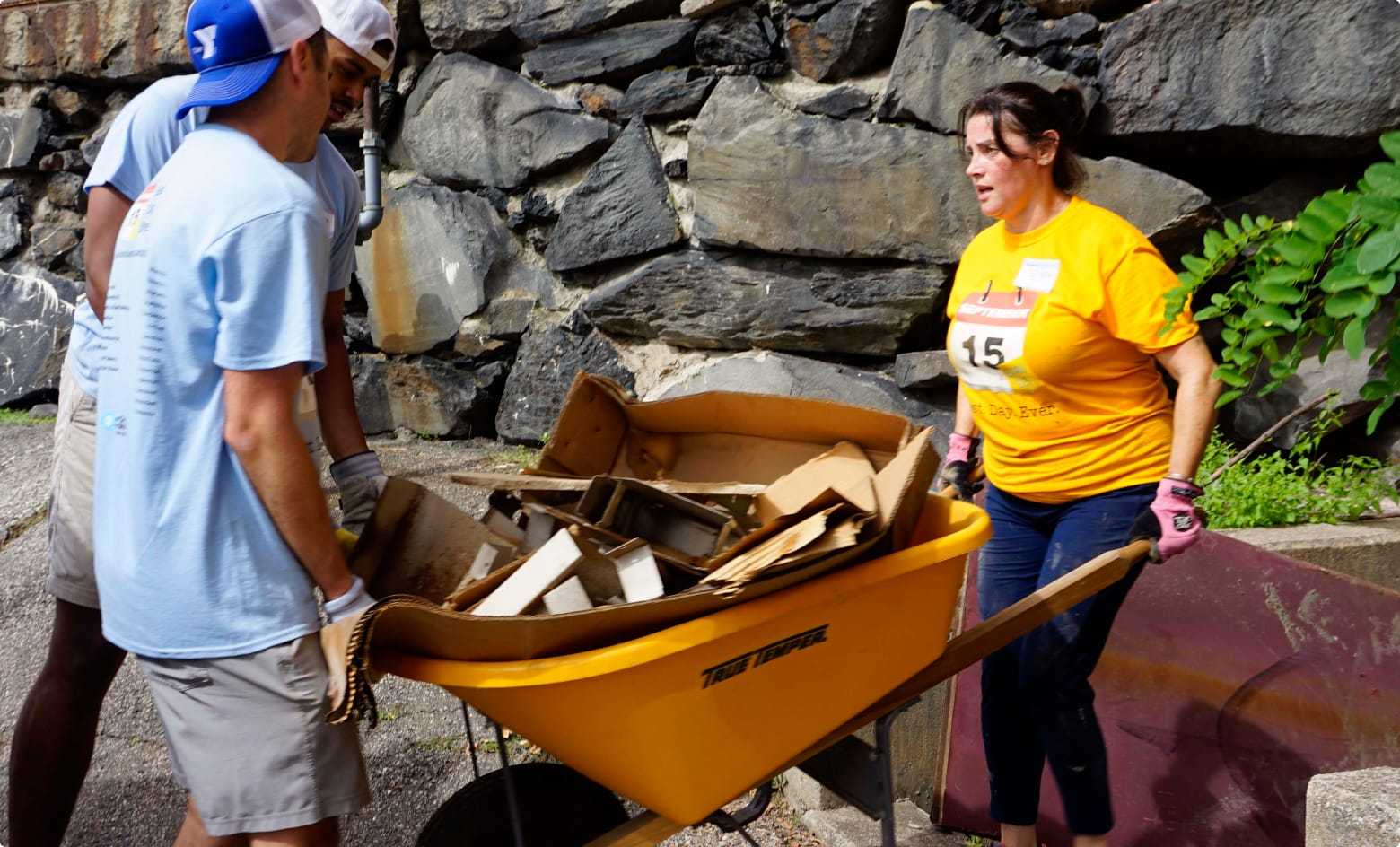 A woman holding a full wheelbarrow while a man helps keep it from tipping over.